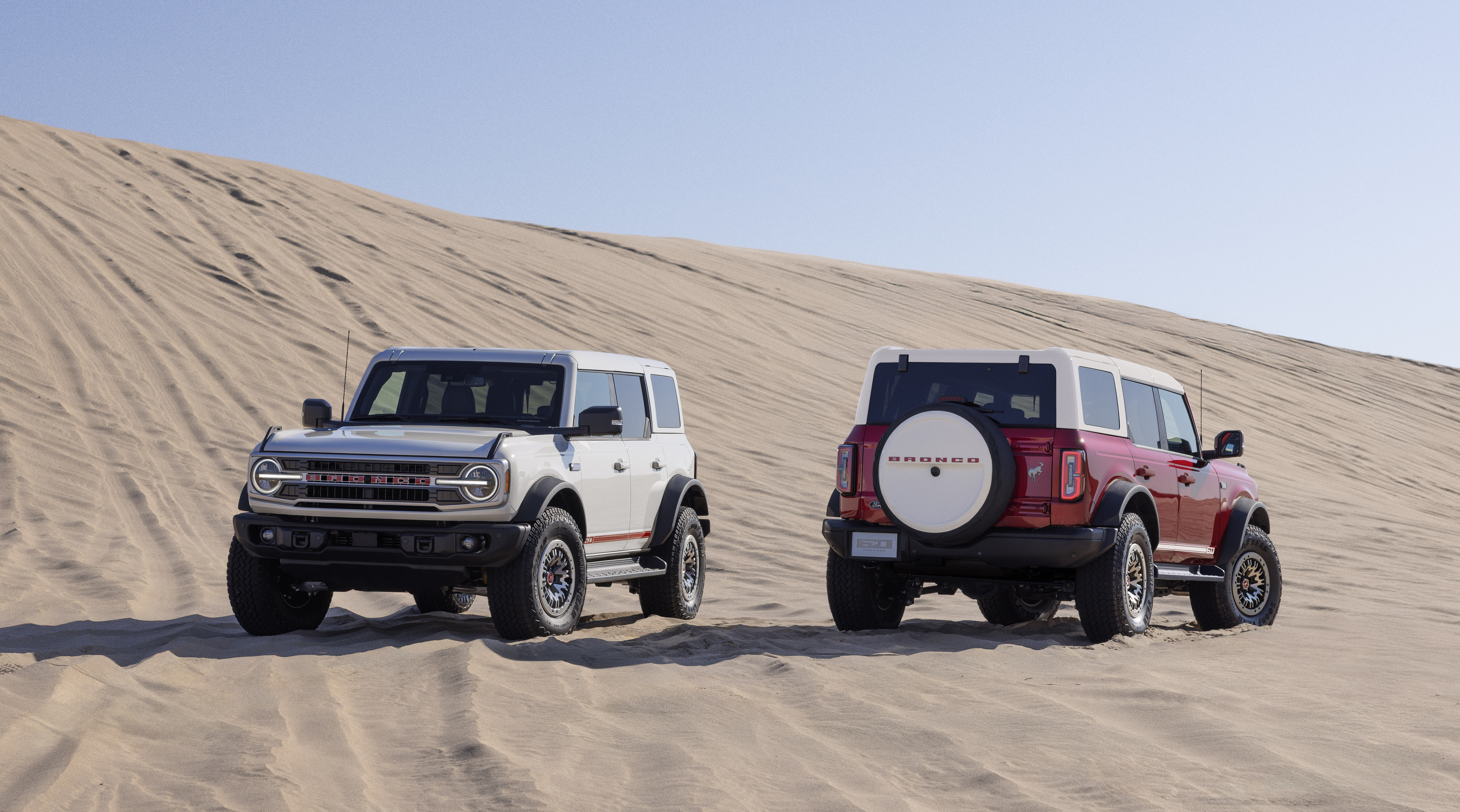 2026 Ford Bronco 60th Anniversary Package shown on two Broncos, one white with red accents and one red with white accents. Vehicles are parked on a sandy hill with a blue sky overhead.